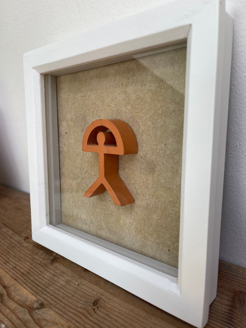 May include: A framed orange-colored wooden sculpture of a stylized human figure with a rounded top, resembling a head, and a curved line extending from the head, resembling an umbrella. The sculpture is mounted on a beige background.