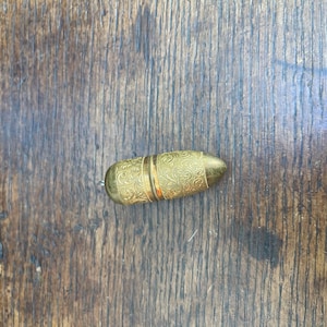 Könnte beinhalten: Eine kleine, goldfarbene Pillendose mit einem verzierten, gravierten Blumenmuster. Die Dose ist oval geformt mit einem Klappdeckel und einem Band in der Mitte. Der Hintergrund ist eine dunkle Holzoberfläche.