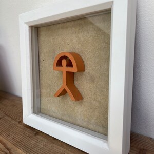 May include: A framed orange-colored abstract sculpture of a person with a rounded top, resembling a head, and a wide base, resembling legs. The sculpture is set against a beige background.