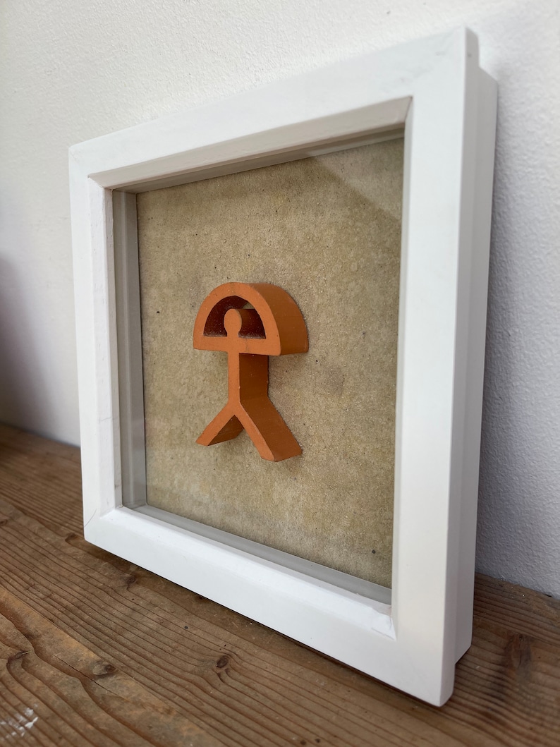May include: A framed orange-colored abstract sculpture of a person with a rounded top, resembling a head, and a wide base, resembling legs. The sculpture is set against a beige background.