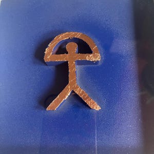 May include: A copper-colored stylized human figure with a rounded arch above its head, mounted on a dark blue background.