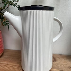 Puede incluir: Tetera de cerámica blanca con tapa negra y diseño acanalado. La tetera tiene un pico y una asa.