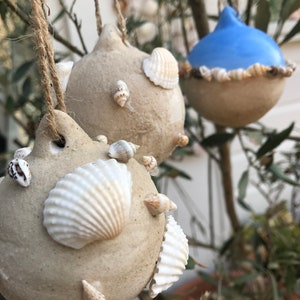 May include: Two beige ceramic ornaments with seashells attached, hanging from a tree branch. One ornament is shaped like a teardrop and the other is round. A blue ceramic ornament with seashells is also hanging from the branch.