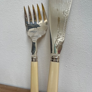 May include: A set of two silver serving utensils with ivory handles. The knife has a decorative design on the blade and the fork has a decorative design on the handle.