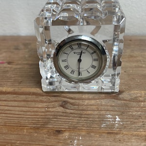 Puede incluir: Un reloj de escritorio de cristal transparente con un diseño cuadrado y facetado. La esfera del reloj es blanca con números romanos y manecillas negras, y las palabras "WATERFORD CRYSTAL QUARTZ" están impresas en la esfera. El reloj está sobre una superficie de madera.