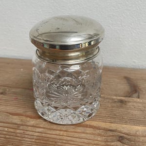 Puede incluir: Un tarro de cristal transparente con tapa plateada. El tarro tiene un diseño tallado y la tapa es de forma simple y redondeada. El tarro está sobre una superficie de madera, con una pared blanca de fondo.