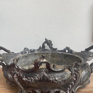 May include: An antique silver-toned decorative bowl with ornate floral and scrollwork detailing. The bowl has a rectangular shape with curved sides, handles, and decorative feet. The interior shows signs of age and use.