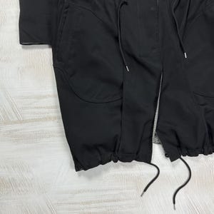 May include: A black jacket featuring a central zipper closure, a snap-button placket, and two large front pockets. The garment includes a drawstring at the waist and a gathered, elasticized hem with additional drawstring cords for adjustment.