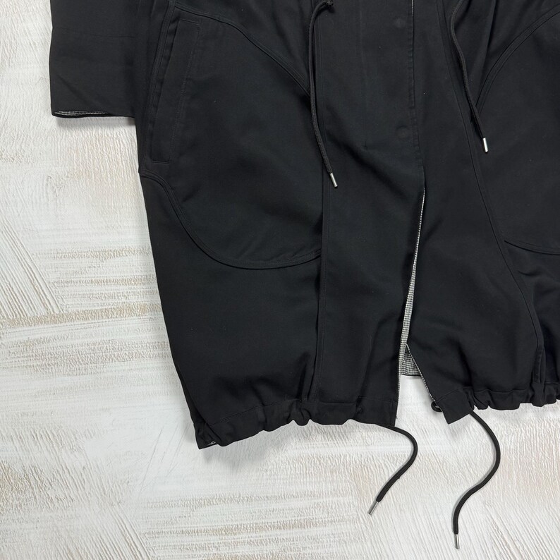 May include: A black jacket featuring a central zipper closure, a snap-button placket, and two large front pockets. The garment includes a drawstring at the waist and a gathered, elasticized hem with additional drawstring cords for adjustment.