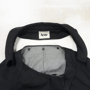 May include: A black jacket with a wide collar and a white interior label featuring the text Acne. The garment has a black and white micro-checkered lining visible at the neckline, secured with three dark metal snap buttons along the upper edge.