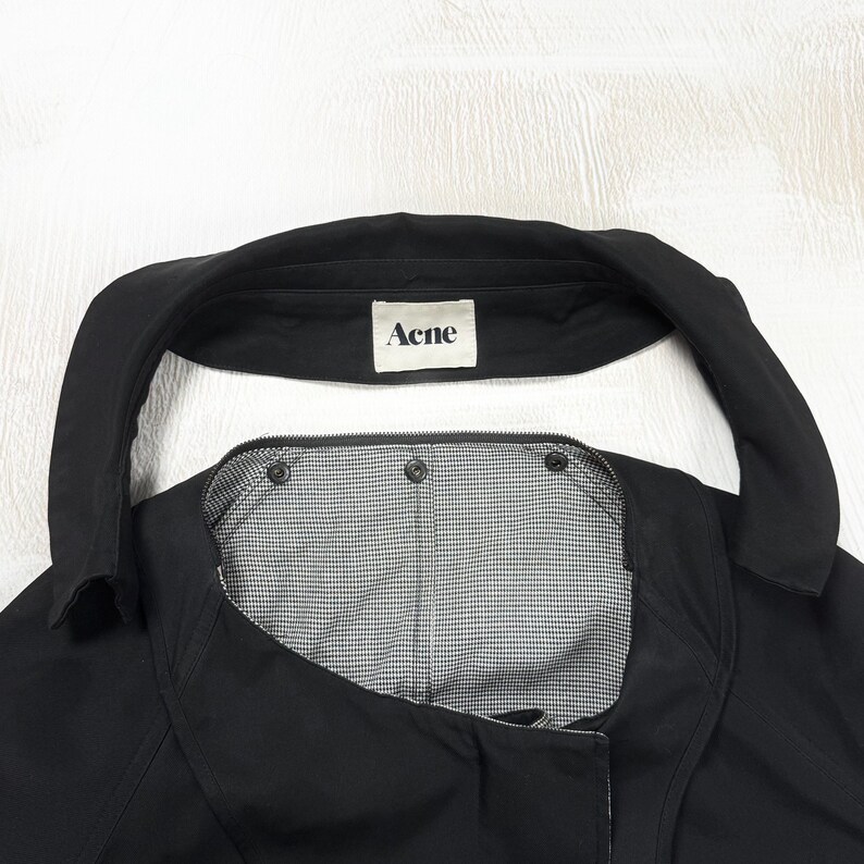 May include: A black jacket with a wide collar and a white interior label featuring the text Acne. The garment has a black and white micro-checkered lining visible at the neckline, secured with three dark metal snap buttons along the upper edge.