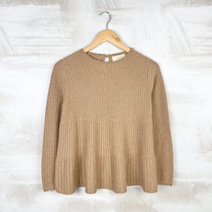 May include: A tan-colored, ribbed knit sweater with long sleeves and a flared hem. The sweater is hanging on a wooden hanger against a white background. The back of the sweater has a small keyhole opening near the neckline.