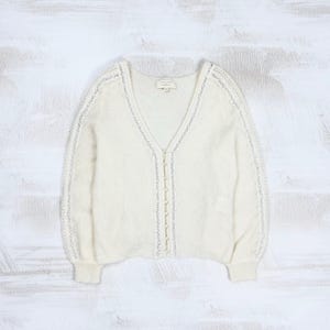 May include: A cream-colored V-neck cardigan sweater featuring long sleeves with intricate white lace trim detailing along the front button placket and down the length of each sleeve. The garment includes a series of small matching fabric buttons.