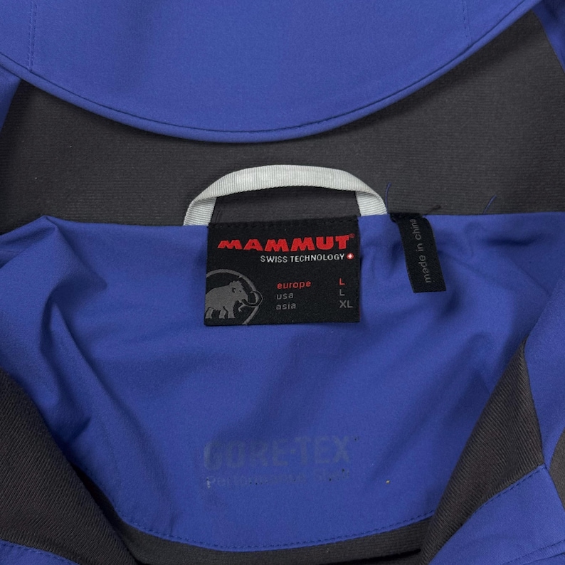 Op de afbeelding: Een blauwe en donkergrijze jas met een zwart binnenlabel met het Mammut-logo, een mammoetafbeelding en tekst die de maten L voor Europa en de VS en XL voor Azi&euml; aangeeft. Het label toont ook de tekst Swiss Technology en Gore-Tex.