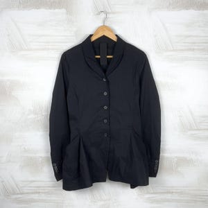 May include: Black button-down jacket with a tailored fit. The jacket features a notched collar, long sleeves with buttoned cuffs, and a pleated design at the waist. It is hanging on a wooden hanger against a white and gray background.