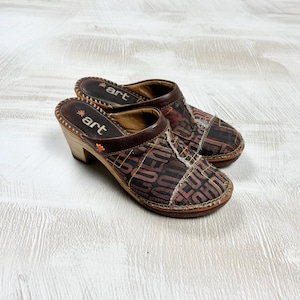 May include: A pair of brown clogs with a wooden heel and a patterned fabric upper. The clogs have a leather trim and the word "art" is visible on the insole. The fabric upper features a repeating pattern in shades of brown and tan.