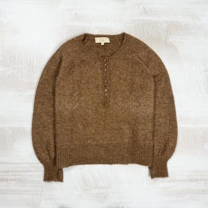 May include: A brown long-sleeve sweater with a round neckline and a vertical placket featuring a row of small buttons. The garment has a textured knit finish, raglan sleeves, and ribbed cuffs and hem. A small rectangular label is at the neck.