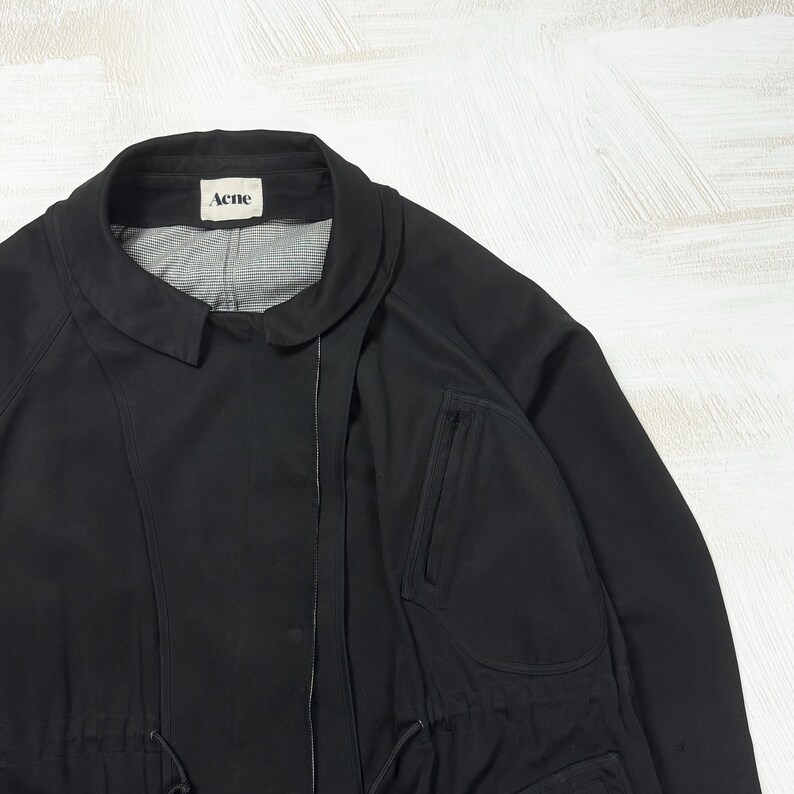 May include: A black jacket featuring a rounded collar, a front zipper closure, and a drawstring waist. The interior lining displays a subtle grey and white checkered pattern. A white label with the black text Acne is sewn into the inner neckline.