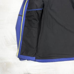 Op de afbeelding: Een blauwe jas met een zwarte binnenvoering en een zwart gaasvak aan de binnenzijde. De jas is voorzien van een felgele ritsrand langs de opening aan de voorzijde, wat zorgt voor een duidelijk kleurcontrast met de blauwe en zwarte stoffen panelen.