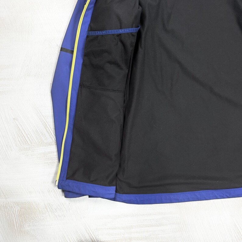 Op de afbeelding: Een blauwe jas met een zwarte binnenvoering en een zwart gaasvak aan de binnenzijde. De jas is voorzien van een felgele ritsrand langs de opening aan de voorzijde, wat zorgt voor een duidelijk kleurcontrast met de blauwe en zwarte stoffen panelen.