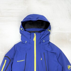 Op de afbeelding: Een koningsblauwe jas met capuchon, voorzien van een felgele centrale ritssluiting en bijpassende gele ritstrekkers op de zakken. De merknaam Mammut en een geel mammoetlogo zijn in contrasterend geel op de borst en mouw gedrukt.
