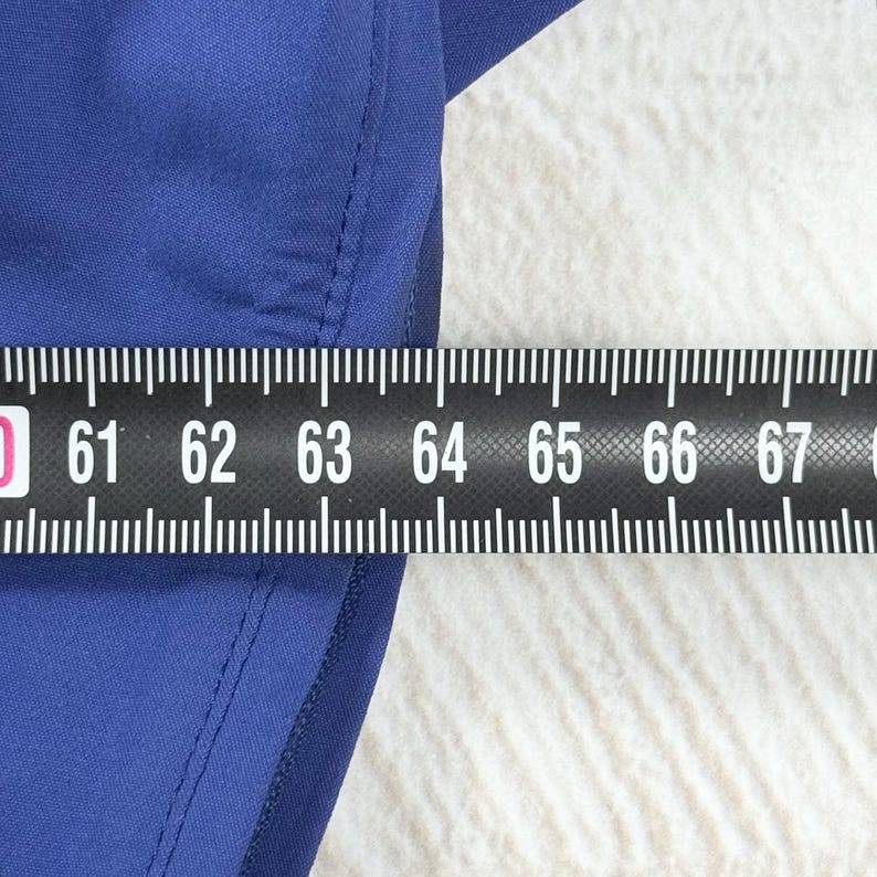 Op de afbeelding: Een effen blauw kledingstuk met een zichtbare naad langs de rand. Een zwart meetlint is over het materiaal geplaatst en toont een meting van 61 tot 67 centimeter, wat de breedte van het blauwe textielstuk aangeeft.