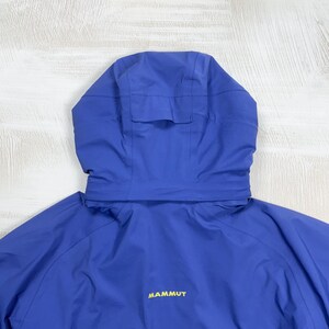 Op de afbeelding: Een felblauw jack met capuchon, voorzien van een gestructureerde capuchon met een verstelbaar lipje aan de achterkant. Op de achterkant van het jack staat het woord MAMMUT in gele letters gedrukt. Het kledingstuk is gemaakt van gladde stof.