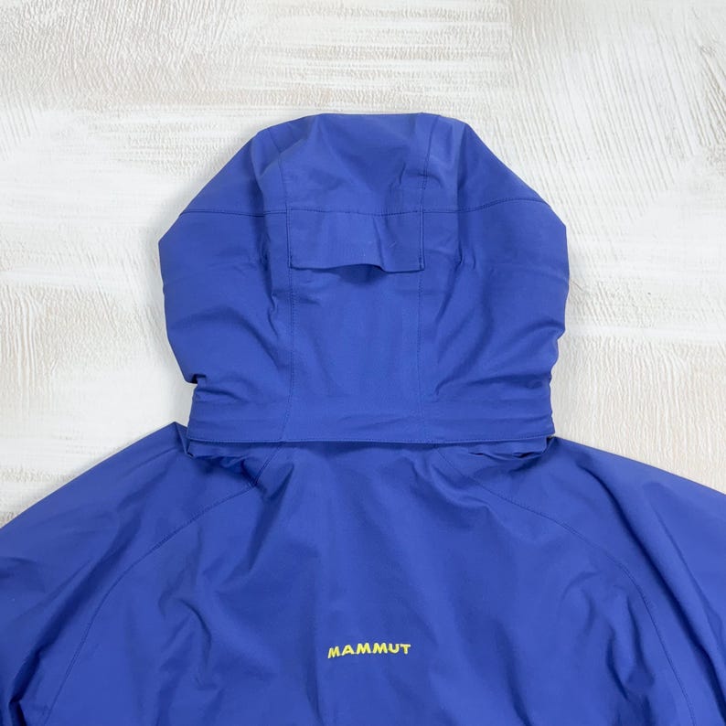 Op de afbeelding: Een felblauw jack met capuchon, voorzien van een gestructureerde capuchon met een verstelbaar lipje aan de achterkant. Op de achterkant van het jack staat het woord MAMMUT in gele letters gedrukt. Het kledingstuk is gemaakt van gladde stof.