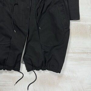May include: A black jacket featuring a front zipper closure, two large side pockets, and a drawstring hem with long black cords. The garment includes a vertical seam detail along the front panels and a visible inner lining with a subtle pattern.