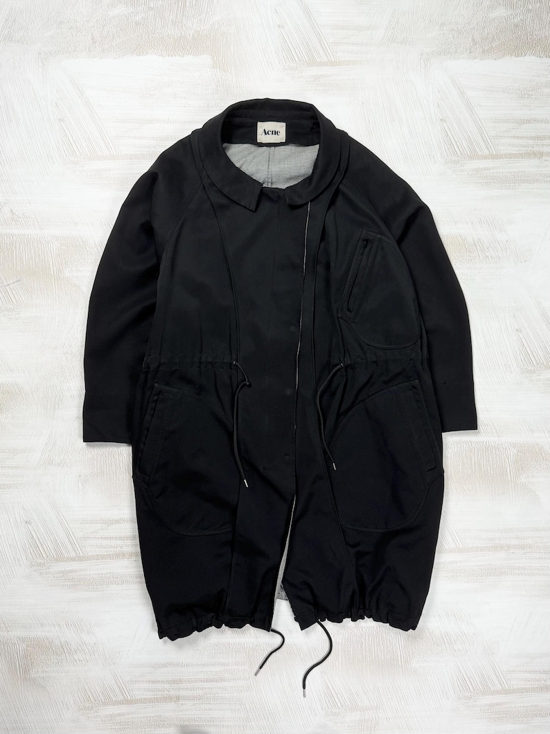 women's black polyamide coat featuring an oversized cyber techwear parka silhouette with utility drawstrings. The structured technical longline jacket with a collared neckline and asymmetrical zipper is displayed flat against a studio background, styled for a fluid cocoon fit corresponding to a clean size Large.
