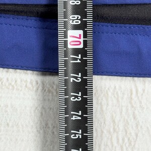 Op de afbeelding: Een blauw kledingstuk met een zwarte rand, geplaatst achter een zwart meetlint dat een meting van ongeveer 70 centimeter aangeeft. De blauwe stof is voorzien van een horizontale naad en een gladde, effen textuur.