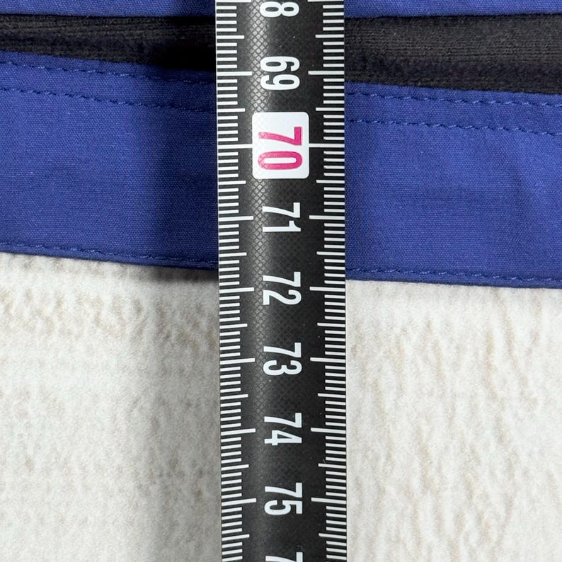 Op de afbeelding: Een blauw kledingstuk met een zwarte rand, geplaatst achter een zwart meetlint dat een meting van ongeveer 70 centimeter aangeeft. De blauwe stof is voorzien van een horizontale naad en een gladde, effen textuur.