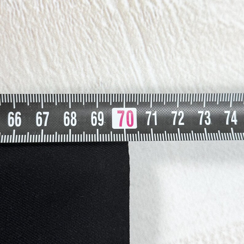 May include: A black fabric edge positioned against a black and white folding ruler. The ruler shows a measurement of 70 centimeters, with the black material aligned precisely at the 70-centimeter mark on the scale for accurate sizing.