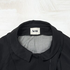 May include: A black jacket with a rounded collar and a white interior lining featuring a small black and white checkered pattern. A white rectangular label is sewn into the inner neckline with the word Acne printed in black capital letters.