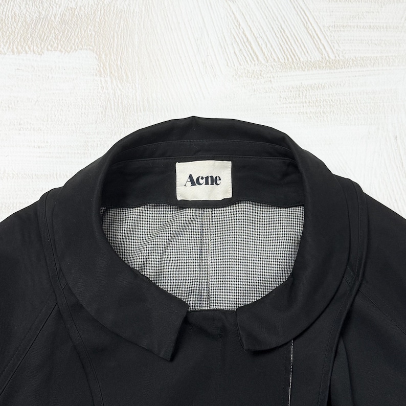 May include: A black jacket with a rounded collar and a white interior lining featuring a small black and white checkered pattern. A white rectangular label is sewn into the inner neckline with the word Acne printed in black capital letters.