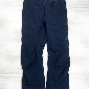 May include: Dark navy blue snow pants featuring a Gore-Tex logo on the right hip and a blue Burton logo on the left leg. These pants include a button and zipper fly closure, belt loops, and zippered pockets on the front thighs and hips.