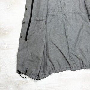 May include: A grey and white pinstriped jacket featuring a black front zipper, a series of black snap buttons along the placket, a large side pocket, and an adjustable drawstring hem at the bottom for a cinched fit and functional design.