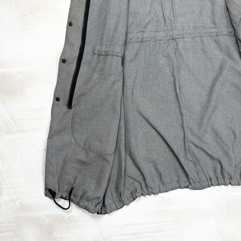 May include: A grey and white pinstriped jacket featuring a black front zipper, a series of black snap buttons along the placket, a large side pocket, and an adjustable drawstring hem at the bottom for a cinched fit and functional design.