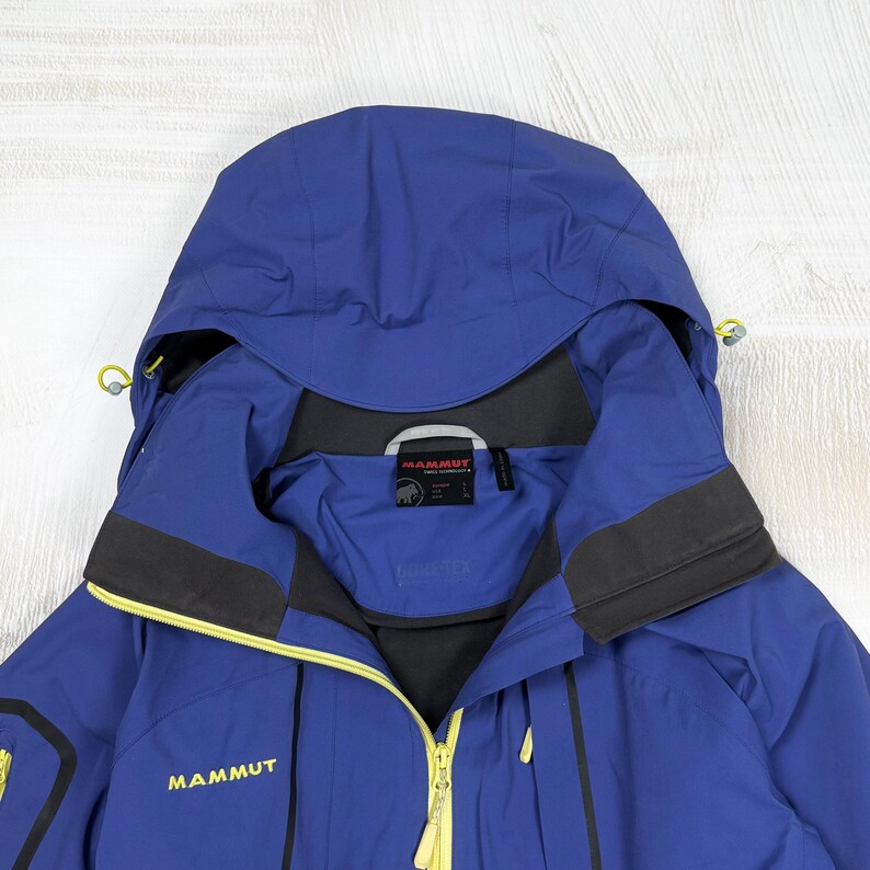 Op de afbeelding: Een blauwe jas met capuchon, voorzien van een gele ritssluiting aan de voorzijde, zwarte accenten en het Mammut-logo in het geel op de borst. De binnenkraag bevat een zwart Mammut-merklabel en een Gore-Tex-logo, met verstelbare trekkoorden.