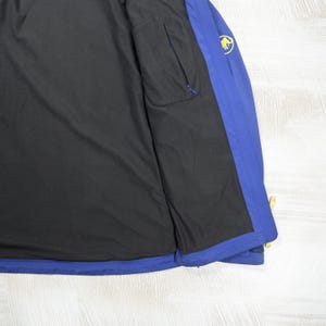 Op de afbeelding: Een blauwe jas met een zwarte binnenvoering. De jas is voorzien van een geel logo van een mammoet op het bovenste gedeelte van de borst en bevat een verticale zak met ritssluiting aan de binnenzijde. De buitenkant is effen blauw van kleur.