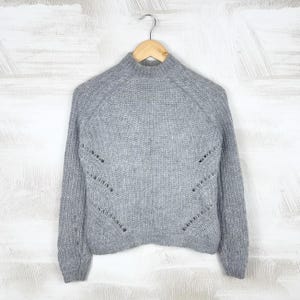 May include: A light gray knit sweater with a mock neck and long sleeves. The sweater has a textured pattern and decorative cut-out details on the front. It is hanging on a wooden hanger against a white background.