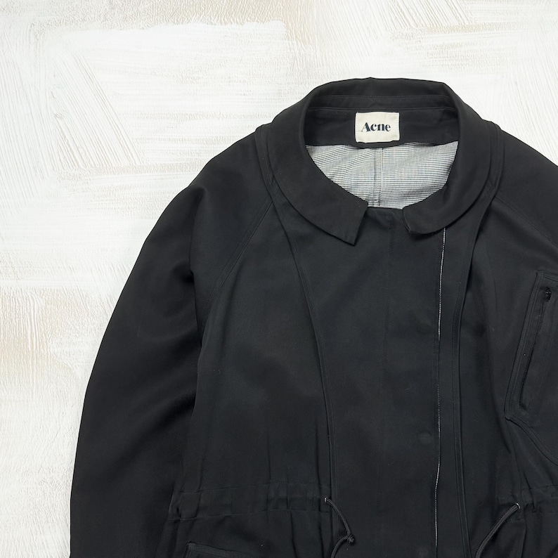 May include: A black jacket with a rounded collar and a white label featuring the text Acne at the neckline. The garment includes a vertical zipper closure, a drawstring waist with visible cords, and a light-colored interior lining with a subtle pattern.