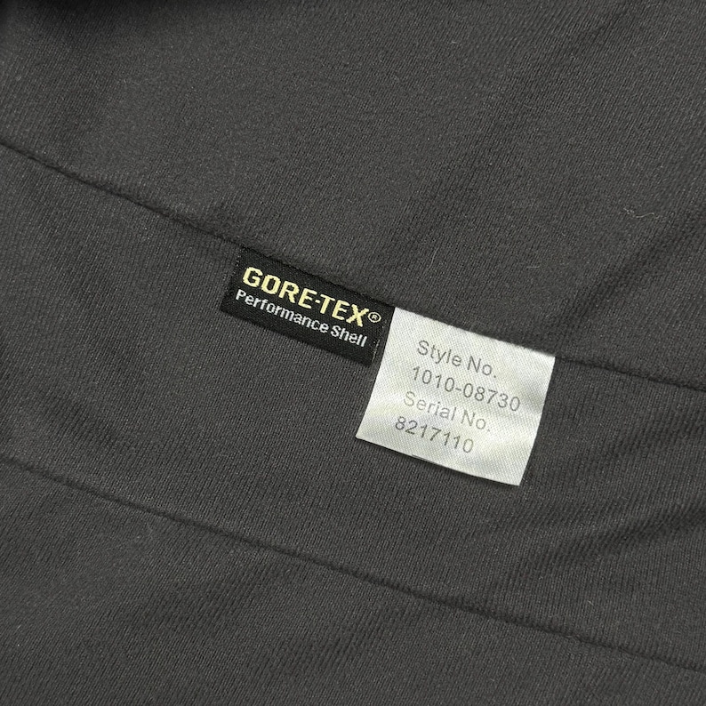 Op de afbeelding: Een donkergrijze stoffen binnenkant met een zwart Gore-Tex Performance Shell label en een wit rechthoekig label met de tekst Style No. 1010-08730 en Serial No. 8217110 in het zwart, met technische details van het kledingstuk.