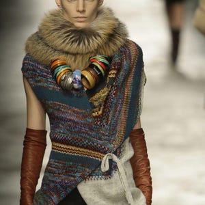 May include: A sleeveless wrap-style knit top featuring a multi-coloured pattern of blue, red, and yellow, paired with a large brown faux fur collar, a chunky necklace of colourful bangles, and long brown leather gloves reaching past the elbows.