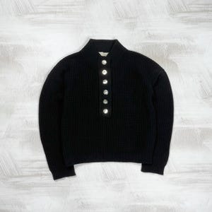 May include: A black long-sleeve sweater featuring a ribbed knit texture, a mock neck collar, and a vertical placket down the center front. The placket is adorned with six round, silver-toned metallic buttons arranged in a single column.