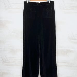 May include: Black velvet pants with two front pockets, hanging on a wooden hanger. The pants have a wide leg design and a button closure at the waist. The fabric has a soft, luxurious sheen.