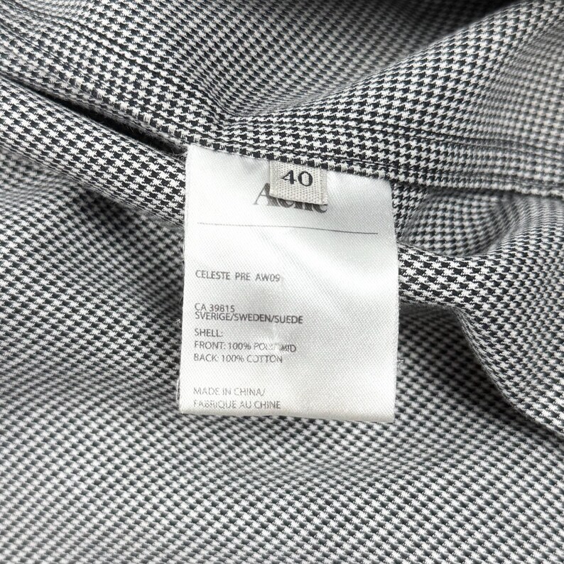 May include: A black and white houndstooth patterned garment with an interior white fabric care label. The label displays the text Acne, size 40, CELESTE PRE AW09, CA 39815, SVERIGE/SWEDEN/SUEDE, SHELL: FRONT: 100% POLYAMID, BACK: 100% COTTON, MADE IN CHINA.