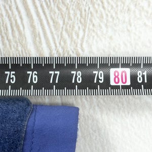 Op de afbeelding: Een blauwe stoffen rand geplaatst onder een zwart meetlint. Het lint toont witte cijfers van 75 tot 81, waarbij het getal 80 is gemarkeerd in een roze vakje, wat een afmeting in centimeters aangeeft voor het blauwe item.