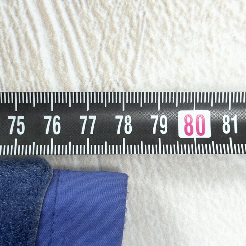 Op de afbeelding: Een blauwe stoffen rand geplaatst onder een zwart meetlint. Het lint toont witte cijfers van 75 tot 81, waarbij het getal 80 is gemarkeerd in een roze vakje, wat een afmeting in centimeters aangeeft voor het blauwe item.