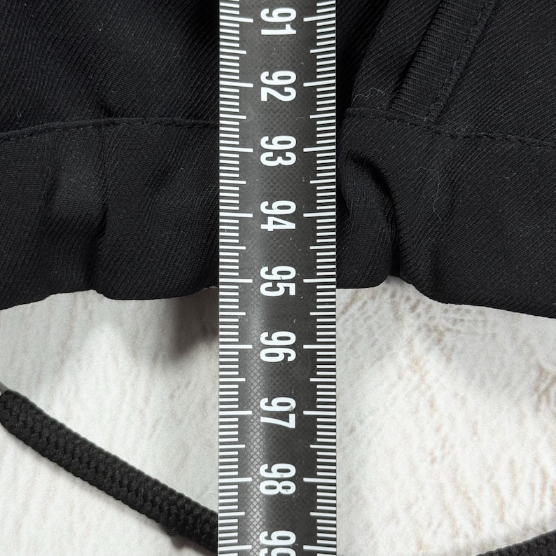 May include: A black garment featuring a drawstring waistband with a black cord, positioned beneath a vertical measuring tape that displays numerical markings from 91 to 99 centimeters, indicating the width of the fabric section shown.